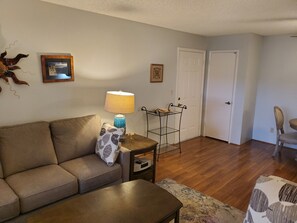 Un salon comprenant un canapé, une table basse, une lampe, une petite table d’appoint et un fauteuil. Il y a deux portes et une étagère fixée au mur, sur laquelle est posé un cadre photo.
