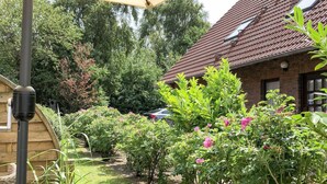 En trädgård med en gångstig som leder fram till ett hus byggt av tegel, omgiven av frodig grönska och blommande växter.