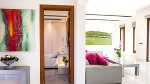Een moderne hotelkamer met een bed, een zithoek met een bank en kussens, uitzicht op een wijngaard en een groot abstract schilderij aan de muur.