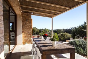 Un patio exterior con una mesa y sillas de madera, rodeado de plantas en macetas y un muro de piedra.