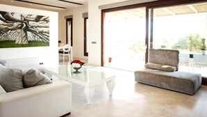 A modern living room with a white sofa, a glass coffee table, and a grey armchair. There is a painting on the wall and a view of the outdoors through large windows.