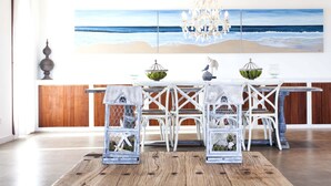 A dining area with a wooden table, white chairs, and a chandelier. There is a large painting of a beach scene on the wall behind the table. - Santolina en Can Corda, Chalet 4 Dormitorios 3 Baños con piscina en Cap de Barbaria Formentera (Sant Francesc de Formentera)