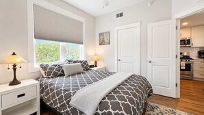 Un dormitorio con cama, mesitas de noche, ventana con persianas, puerta y vistas a una cocina.