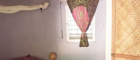 A bedroom with a bed, a window with a curtain, a wooden door, and a woven ceiling.
