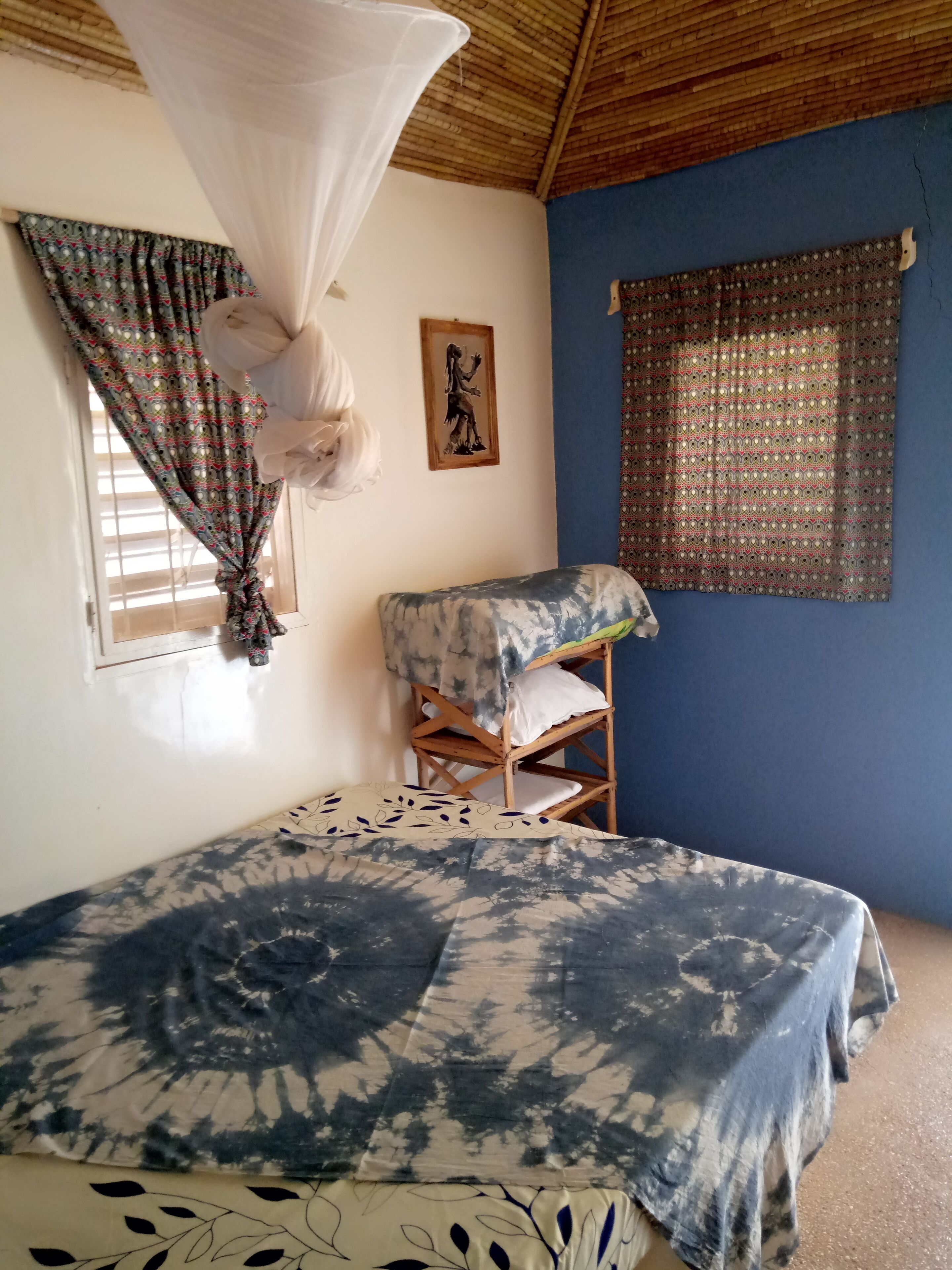 A bedroom with a bed, a wooden chair, a mosquito net, and a window with patterned curtains.