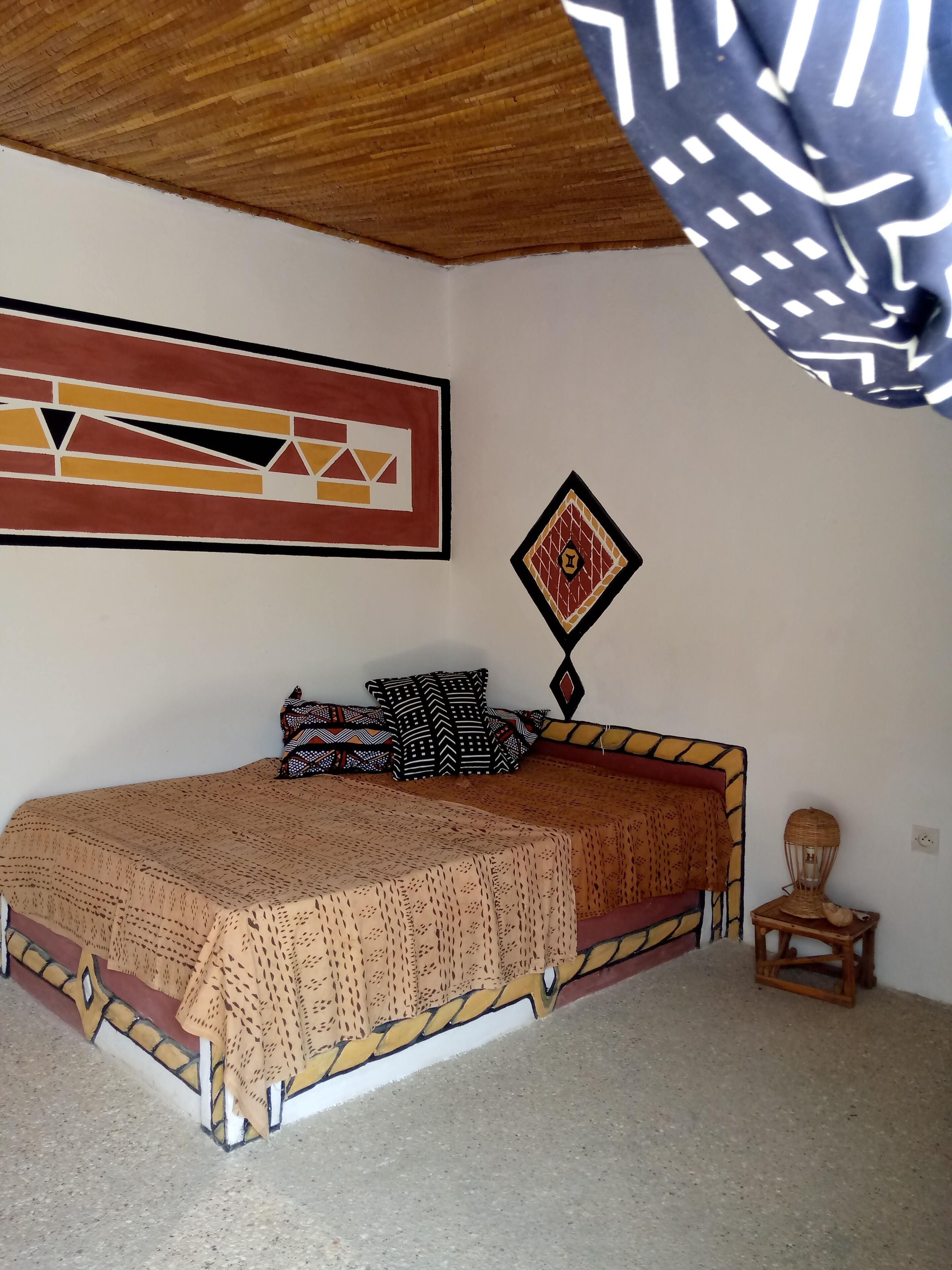 Een kamer met een bed, een muur met geometrische kunst, een decoratief kussen en een kleine tafel met een lamp.