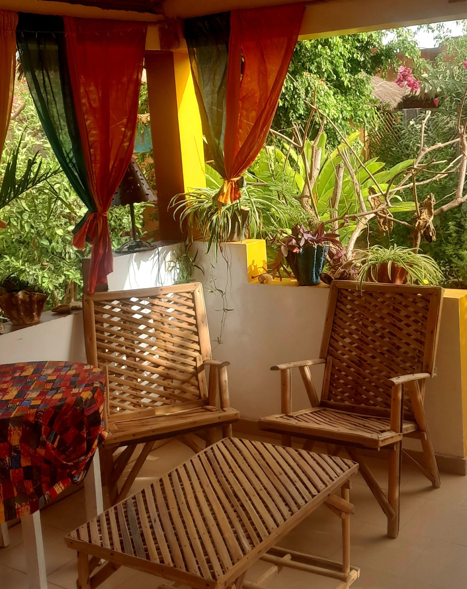 Un espacio al aire libre techado con dos sillas de madera y una mesa, rodeado de plantas en macetas y una pared amarilla.