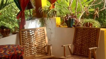 Un espacio al aire libre techado con dos sillas de madera y una mesa, rodeado de plantas en macetas y una pared amarilla.