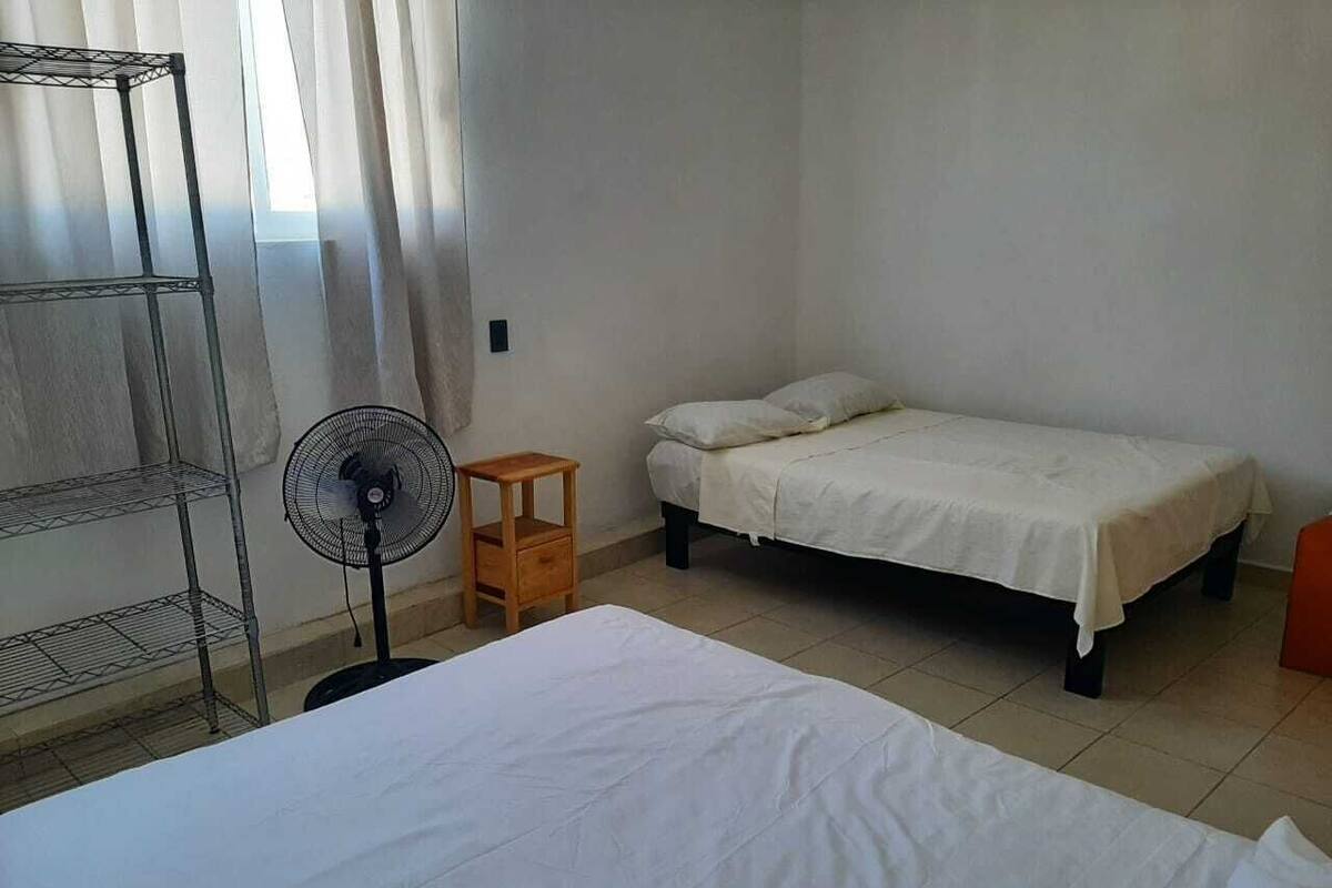 A room with a bed, a small table, a fan, and a shelf unit.