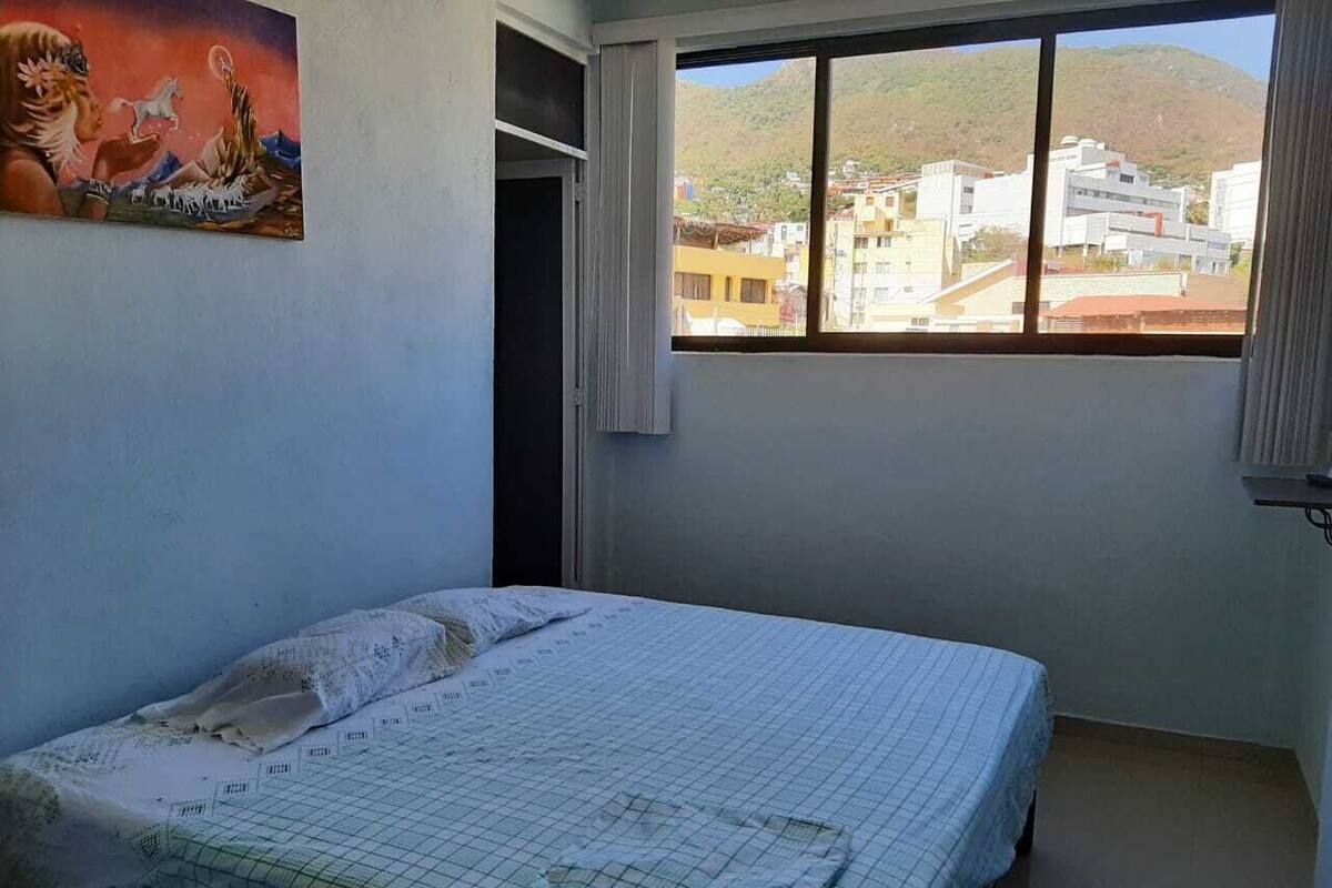 A bedroom with a bed, a window with a view of buildings and hills, and a painting on the wall.