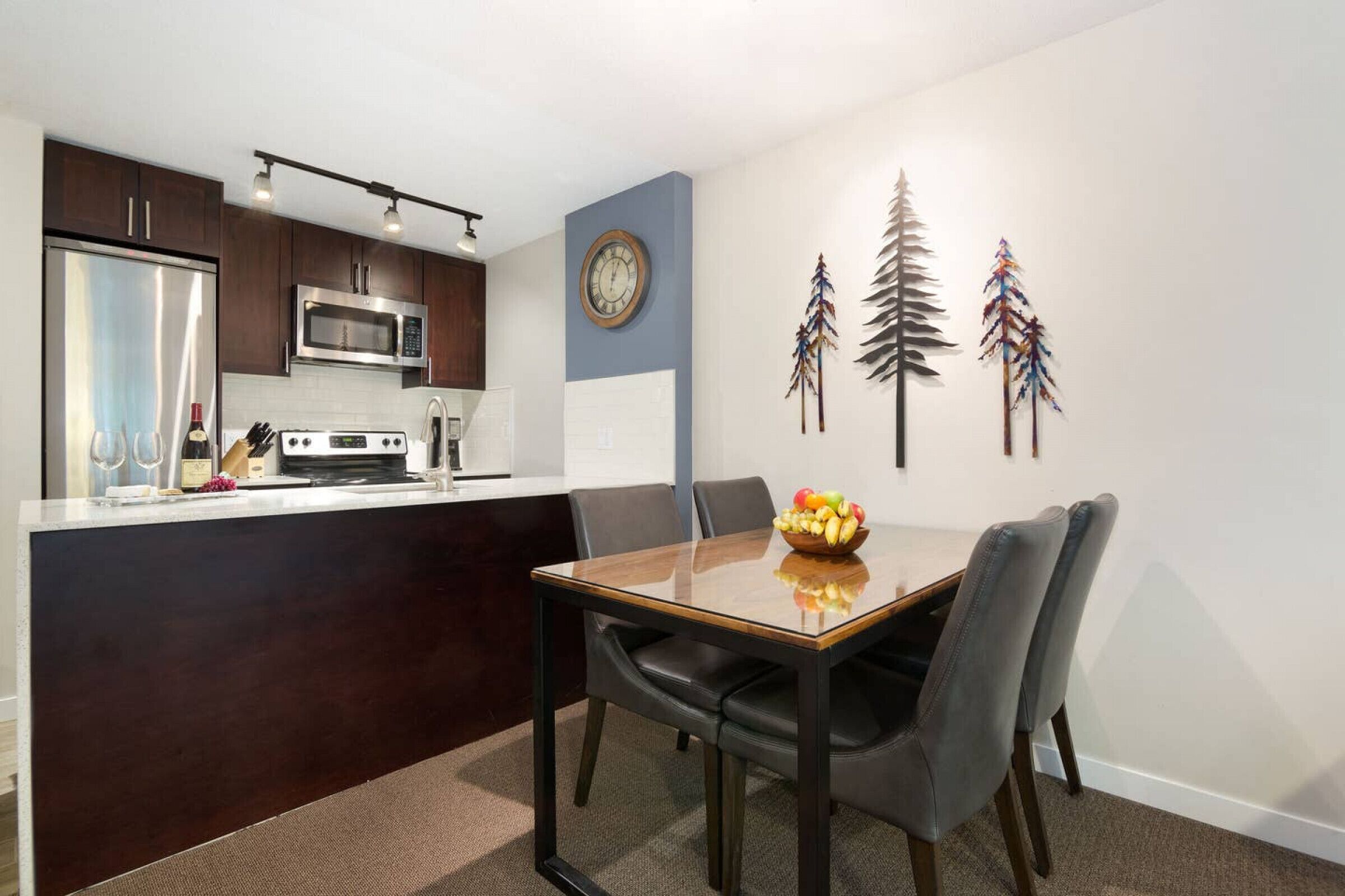 A modern kitchen with dark wood cabinets, a stainless steel refrigerator, and a microwave. There is a dining table with four chairs and a fruit bowl on it. The wall features a clock and decorative wall art.