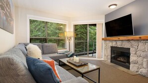 A living room with a stone fireplace, a large flat-screen TV mounted above it, a grey sofa with cushions, a coffee table, and a window with blinds. - Luxury Ski-in/out 1Br/1Ba - 3Hot Tub`s & Pool (Whistler)