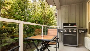 A balcony with a table and chairs, a Coleman grill, and a view of a forest. - Luxury Ski-in/out 1Br/1Ba - 3Hot Tub`s & Pool (Whistler)