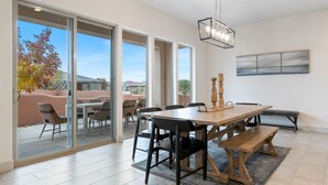 A spacious dining area with a large wooden table, black chairs, and a bench. There is a sliding glass door leading to an outdoor patio with chairs.