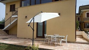 Una casa de dos pisos con un patio que incluye una mesa, sillas, un paraguas blanco y plantas en macetas.