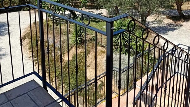 Un balcón con barandilla metálica que da a un jardín con olivos y un cielo azul despejado.