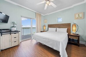 A bedroom with a bed, a TV, a ceiling fan, a nightstand with a lamp, a dresser, and a balcony door.