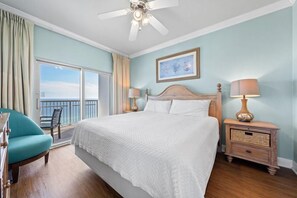A bedroom with a large bed, a ceiling fan, a nightstand with a lamp, a chair, and a window with a view of the sea. - Crystal Tower 1801 (Gulf Shores)
