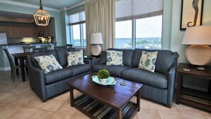 A living room with a sofa set, a coffee table with a plant, a dining area with chairs, a kitchen area, and a large window with a view. - Crystal Tower 1801 (Gulf Shores)