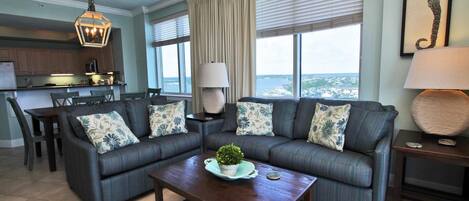 A living room with a sofa set, a coffee table with a plant, a dining area with chairs, a kitchen area, and a large window with a view.