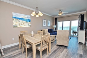 A dining area with a wooden table and chairs, a ceiling fan, and a painting of a beach scene. - Crystal Tower 1702 (Gulf Shores)