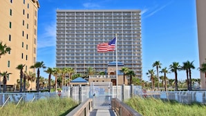 A hotel with a large American flag, a boardwalk leading to the entrance, and palm trees around. - Crystal Tower 1205 by Youngs Suncoast (Gulf Shores)