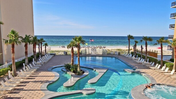A pool area with a view of the beach, lounge chairs, and palm trees. - Crystal Tower 1507 (Gulf Shores)