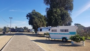 Ein Campingplatz mit Reisemobilen auf einem Kiesplatz, einem gepflasterten Weg und einem großen Baum im Hintergrund.