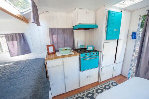 Una kitchenette compacta con estufa azul, armarios blancos y una ventana con cortinas.