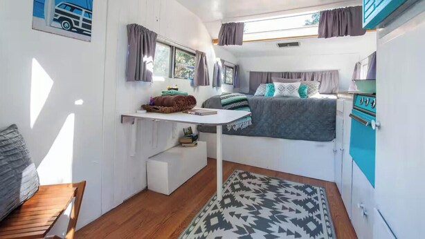 Ein kompaktes Wohnmobil-Interieur mit Bett, kleinem Schreibtisch, Fenster und Küchenzeile.