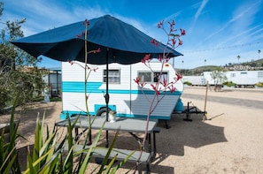 A mobile home with a blue and white exterior, a table with a blue umbrella, and red flowering plants in the foreground.