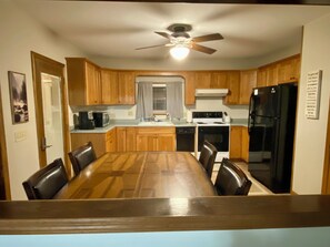 A compact kitchen with wooden cabinets, a dining table with chairs, a refrigerator, a microwave, and a ceiling fan.
