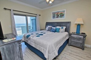 A bedroom with a large bed, a nightstand, a desk, and a view of the ocean through a sliding door.