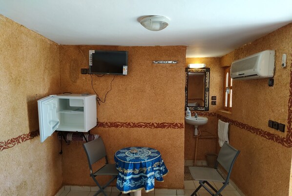 Une pièce de taille modeste, avec un téléviseur fixé au mur, une petite table recouverte d’une nappe à motifs, une chaise, un lavabo et un appareil de climatisation.