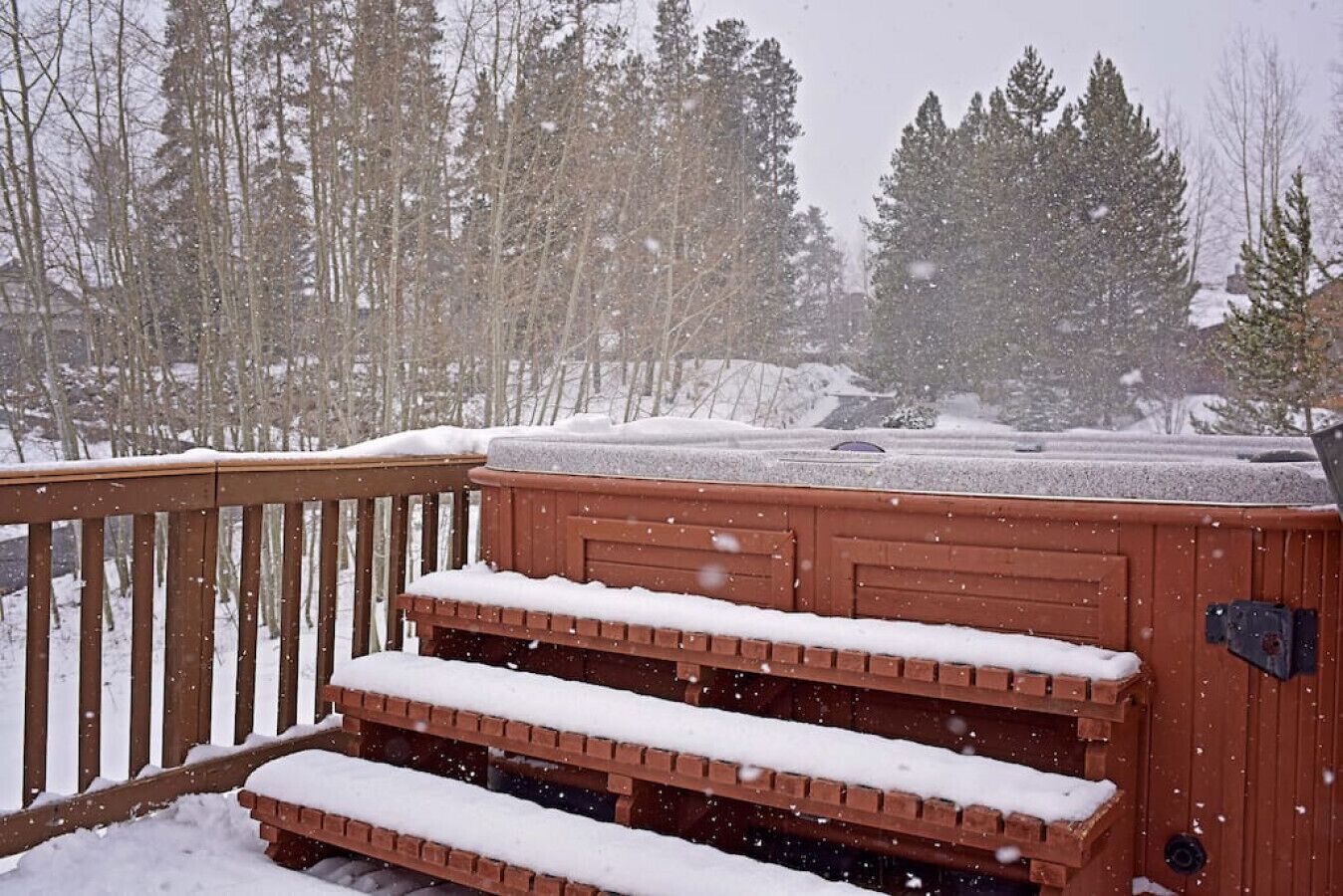 A wooden deck with steps covered in snow, overlooking a snowy landscape with trees.