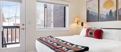 A bedroom with a bed, a window, a door, and a wall-mounted lamp.