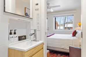 Ein modernes Badezimmer mit Waschbecken, Spiegel und Dusche. Ein Bett mit weißer Bettwäsche und ein Fenster mit Blick auf Bäume und Gebäude.