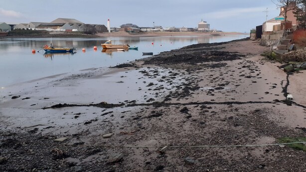 Un paysage côtier avec des bateaux sur une eau calme, une plage de sable et des bâtiments en arrière-plan.