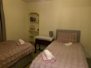 Habitación de hotel con dos camas, cada una con una toalla enrollada. Se observa un mesita de noche con una lámpara y una estantería con libros.