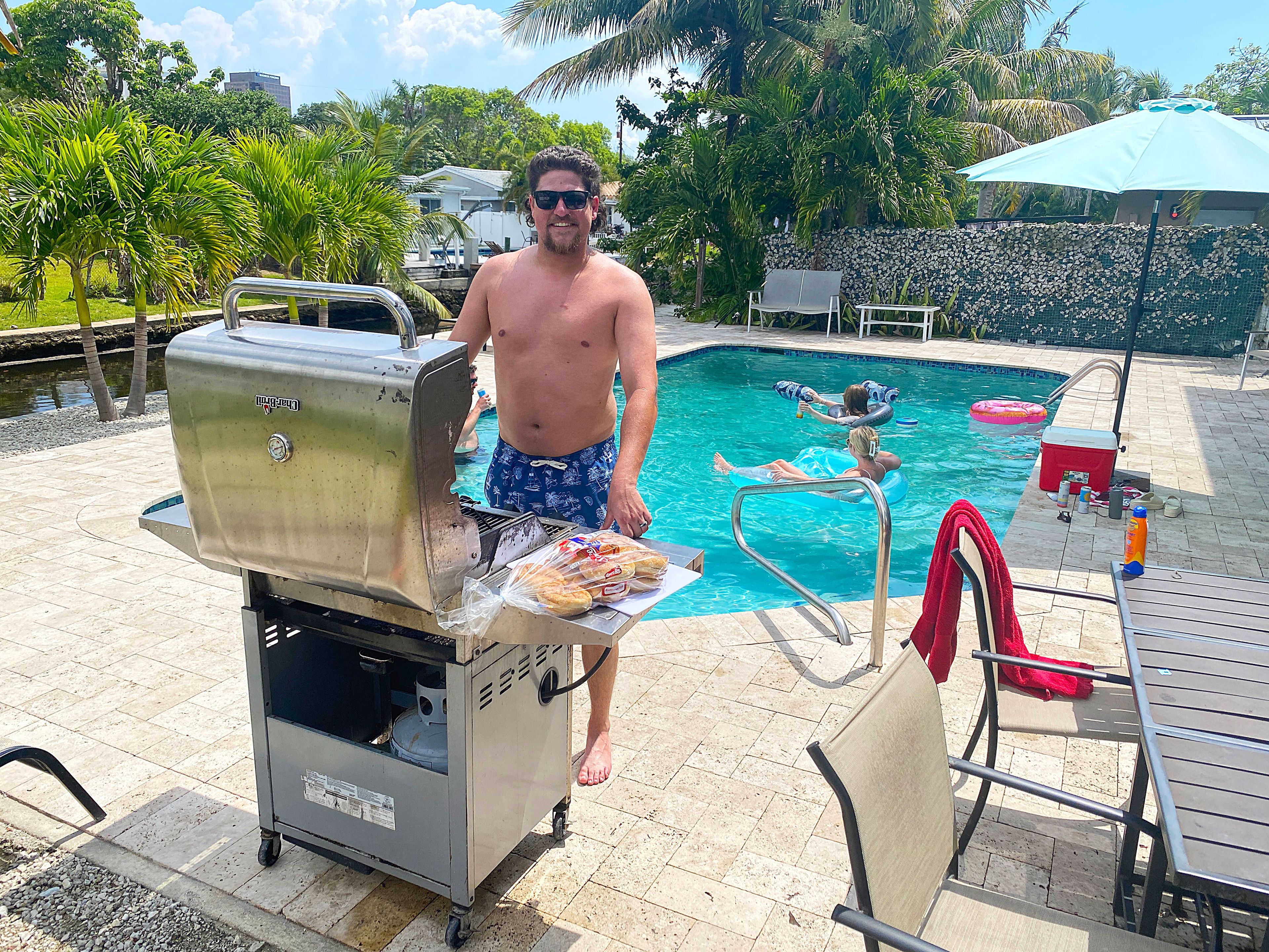 A man grilling outdoors near a swimming pool with lounge chairs and a water feature.