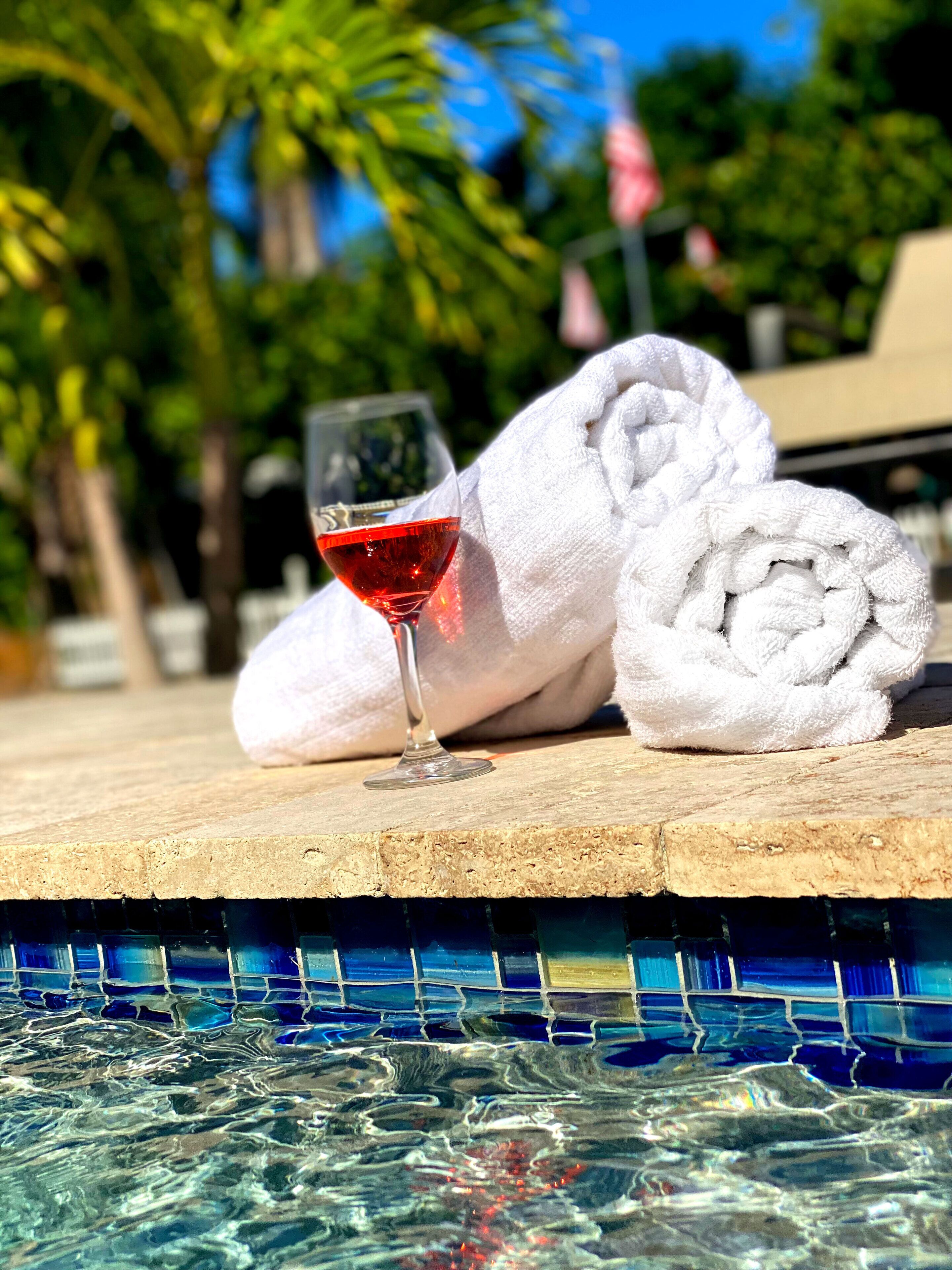 Une scène de bord de piscine avec un verre de vin rouge, une serviette blanche roulée et le bord de la piscine recouvert de carreaux.
