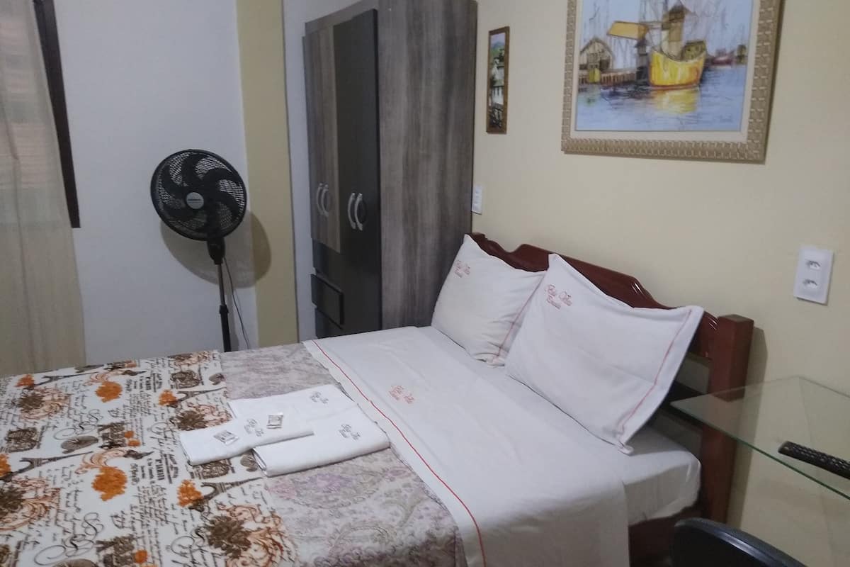 A hotel room with a bed, a fan, a wardrobe, and a painting on the wall.
