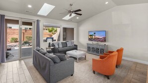 Un salon moderne avec un canapé gris, des fauteuils orange, un téléviseur et un ventilateur de plafond.