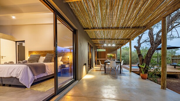 A modern bedroom with a large bed, a dining area with a table and chairs, and a covered outdoor space with a view of trees.