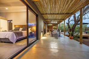 A modern bedroom with a large bed, a dining area with a table and chairs, and a covered outdoor space with a view of trees. - 444 on Taaibos Bush Villa (Hoedspruit)