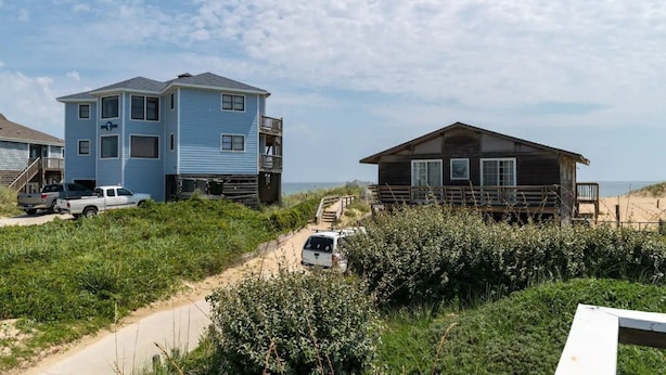 Una casa de playa de dos pisos con un balcón, un sendero arenoso que lleva a la entrada y vista al océano.