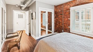 Un dormitorio con pared de ladrillo, un ventanal con contraventanas, una cama y una puerta que da a otra habitación.