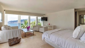 Una camera d'albergo con un letto, due divani bianchi, un tavolino, una TV e vista mare.