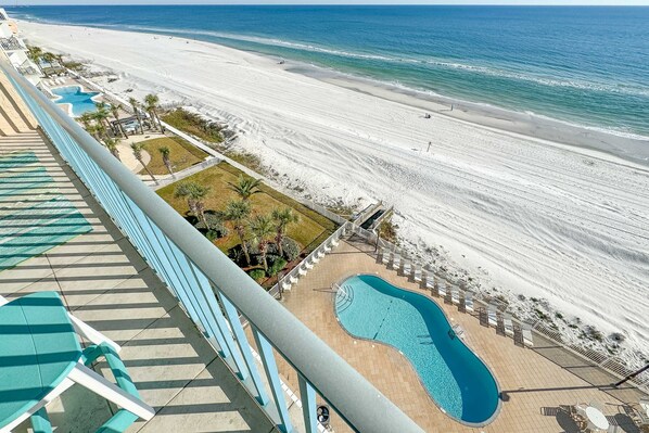 A beach view from a balcony with a swimming pool and lounge chairs.