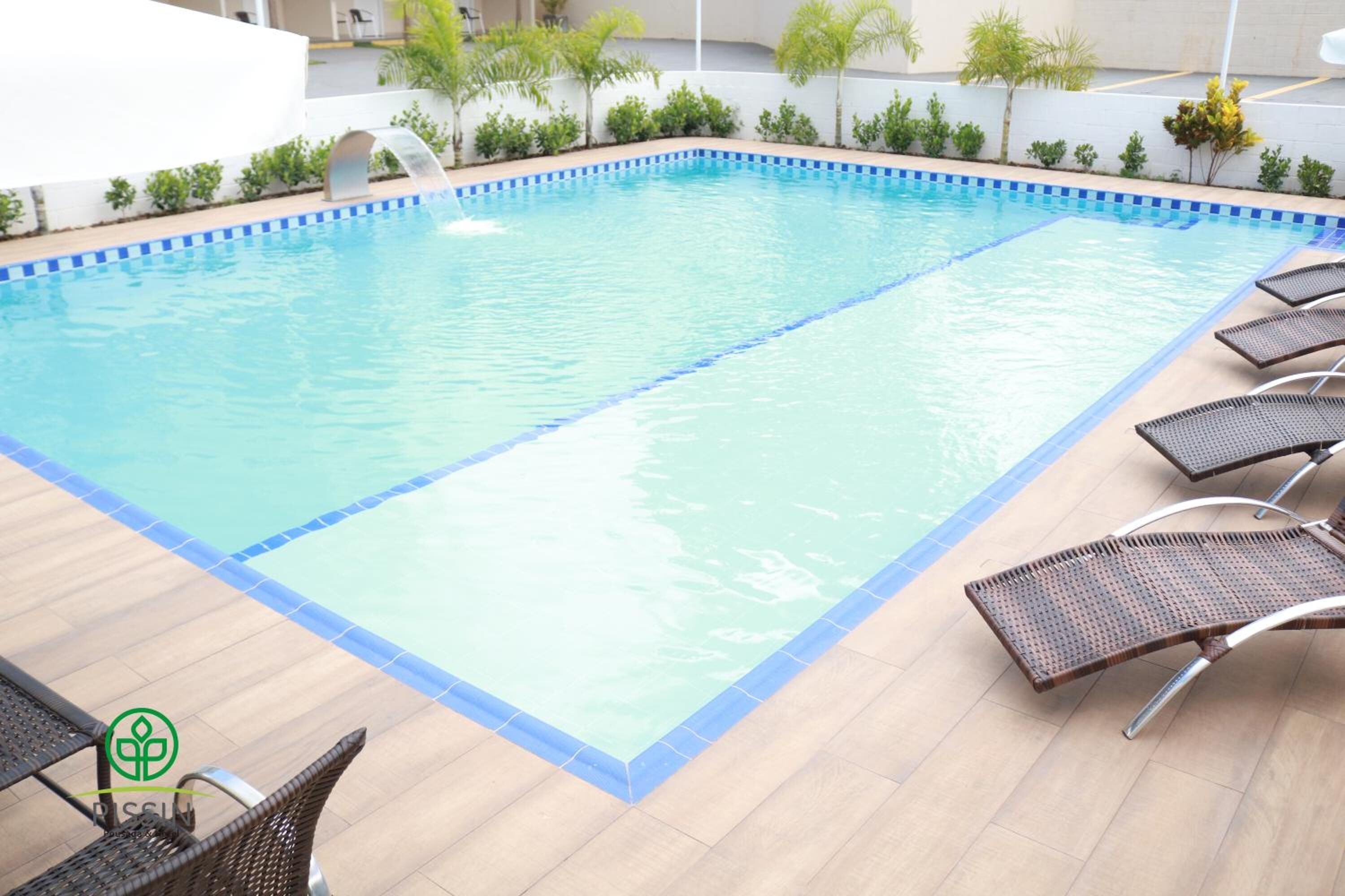 En rektangulær swimmingpool med blå kant, omgivet af en fliseterrasse. Der er liggestole placeret langs poolens kant.
